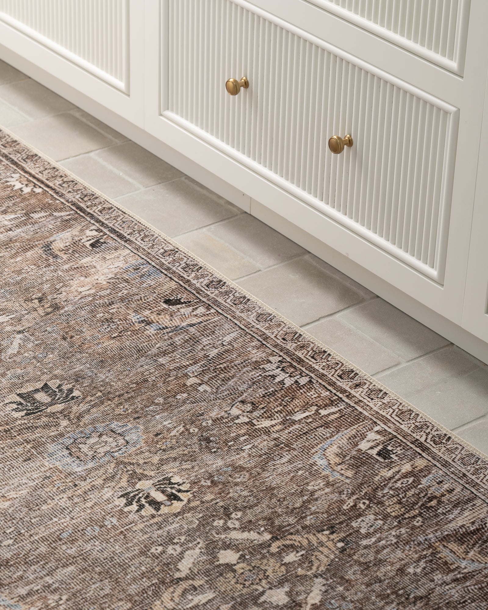 A textured vintage rug with ornate patterns lies on a light beige tile floor beside white cabinetry with gold knobs, creating a cozy, classic ambiance. | SW Home