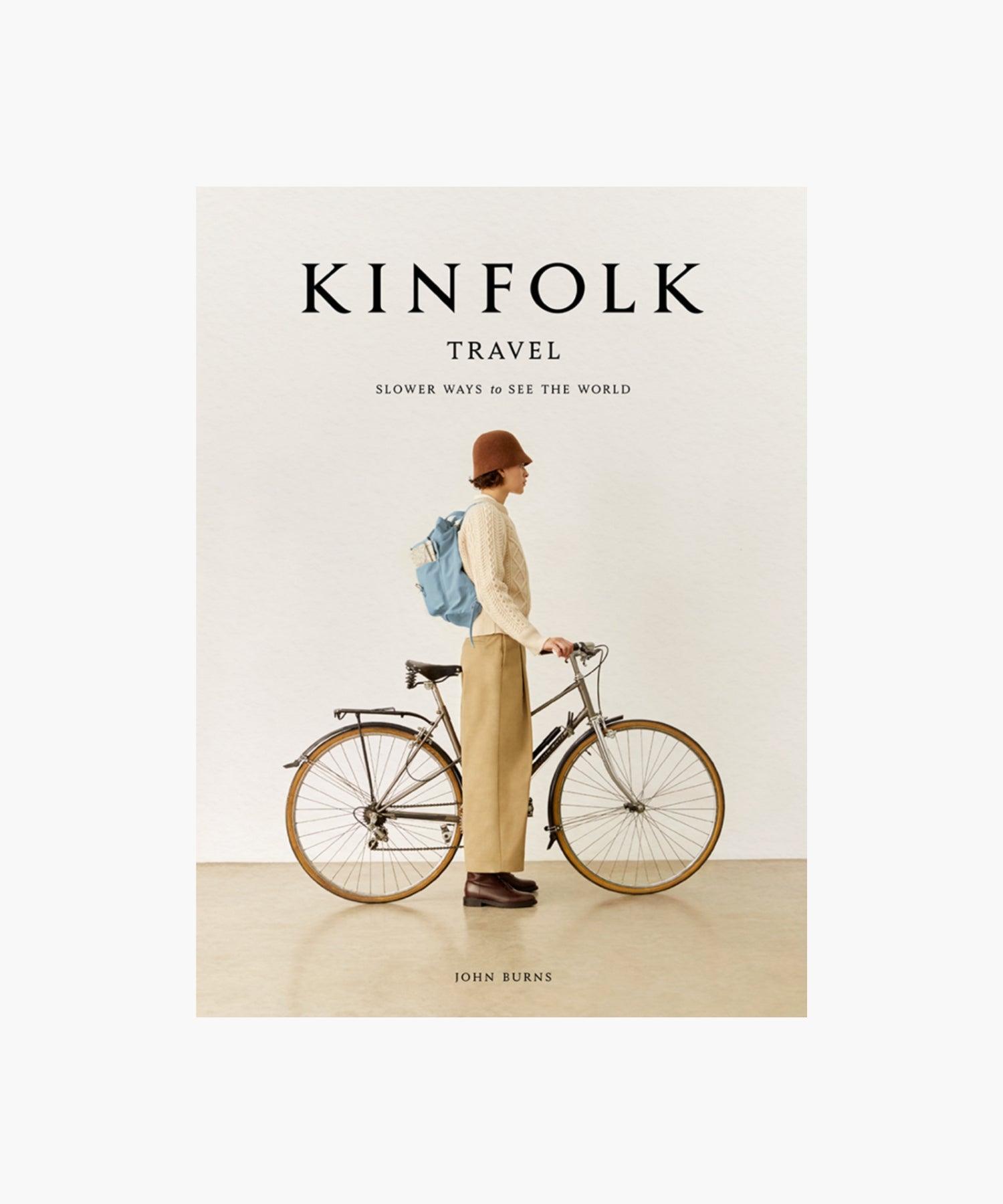The cover of the book "Kinfolk Travel" by John Burns features a side view of a person in a brown hat, beige sweater, and khaki pants, standing next to a bicycle with a blue backpack. The tone is calm and nostalgic. | S.W. Home
