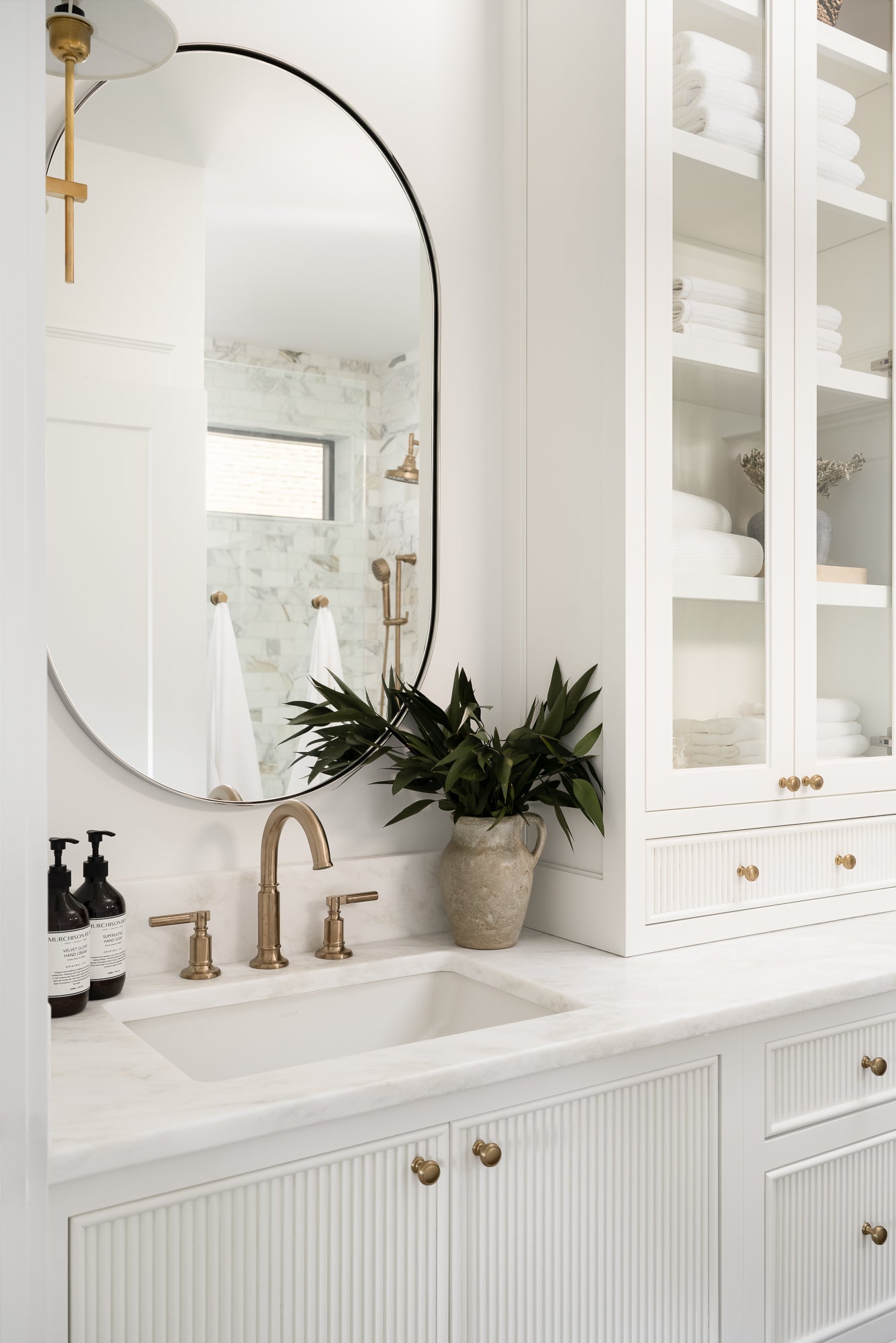 Elegant white bathroom with marble countertop, gold faucets, and oval mirror. Glass cabinet holds towels. Potted plant adds greenery. Bright, serene feel.
