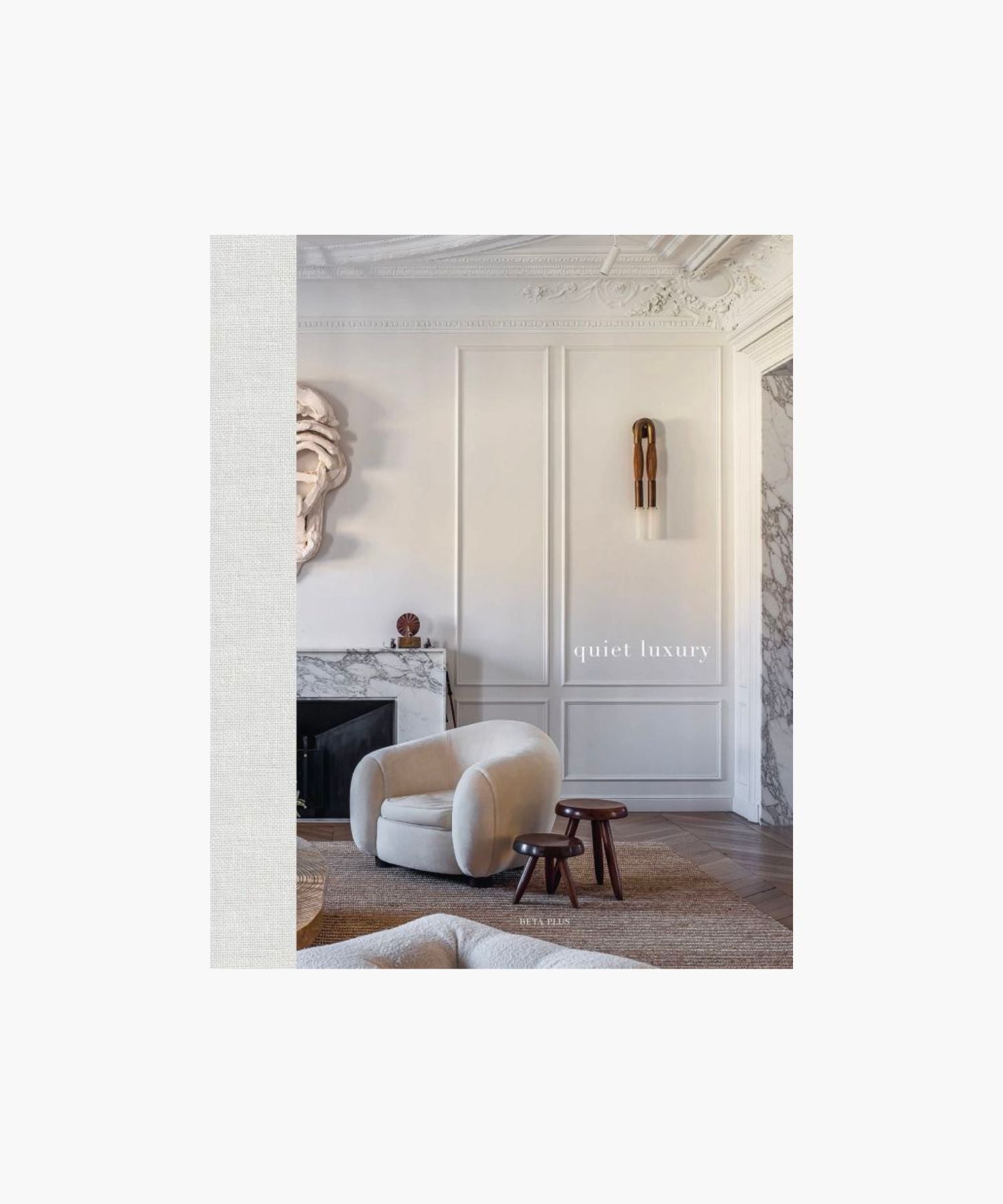 The cover of the book "Quiet Luxury" features a sophisticated living room with a plush beige armchair, two dark wood stools, ornate wall paneling, and a marble fireplace. | S.W. Home