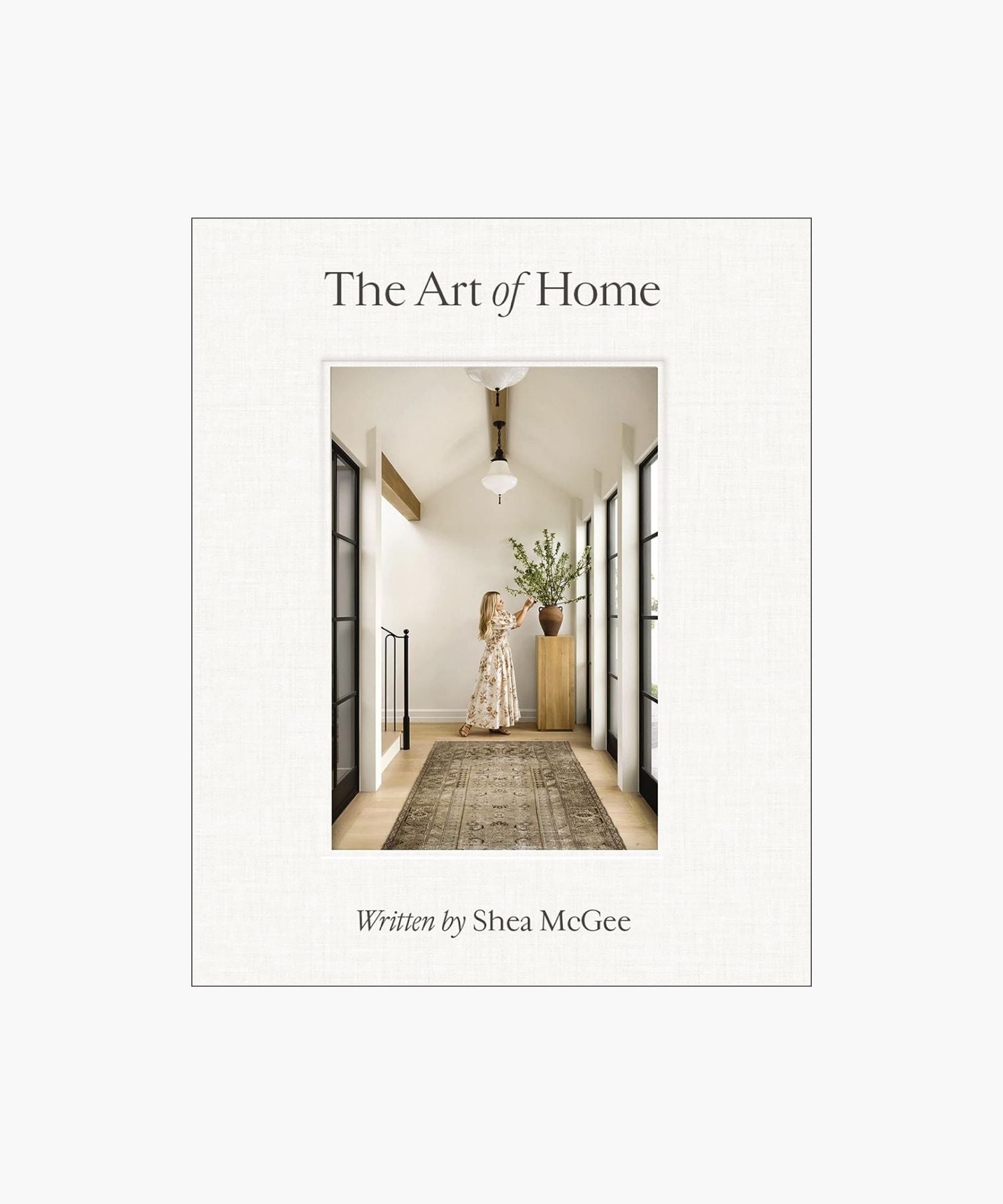 The cover of the book "The Art of Home" by Shea McGee features a woman arranges branches in a vase on a wooden stand, in a bright hallway with large windows and a patterned rug. | S.W. Home
