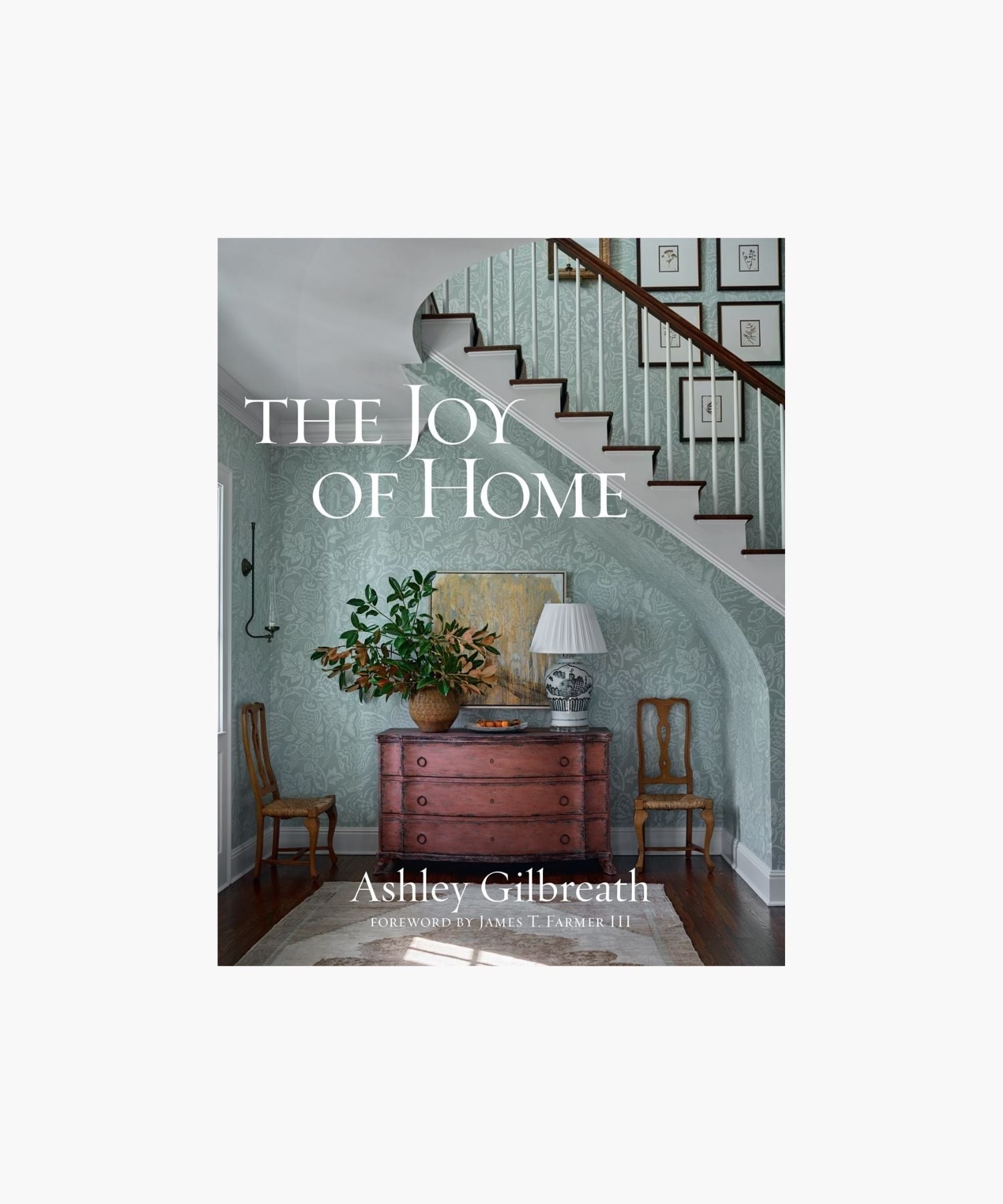 The cover of the book "The Joy of Home" by Ashley Gilbreath features a cozy, elegant hallway with a wooden chest, potted plant, and lamp beneath a staircase; framed prints on the staircase wall. Classic and inviting. | S.W. Home