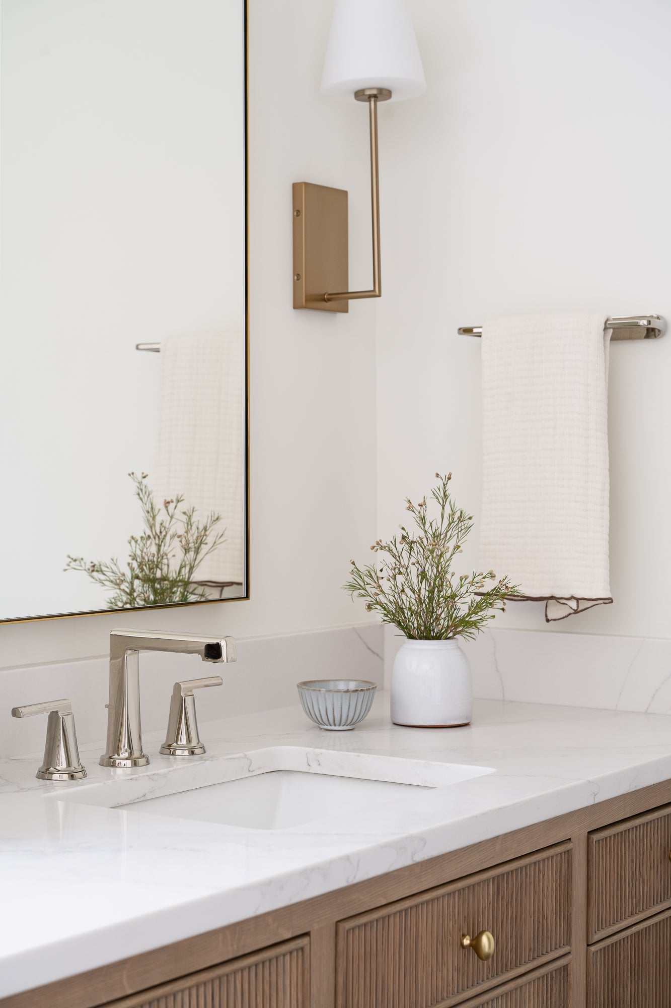 A modern bathroom vanity with a white marble countertop, silver faucet, and wooden cabinets. A towel hangs on the wall, with a vase of greenery and a small bowl adding decor.