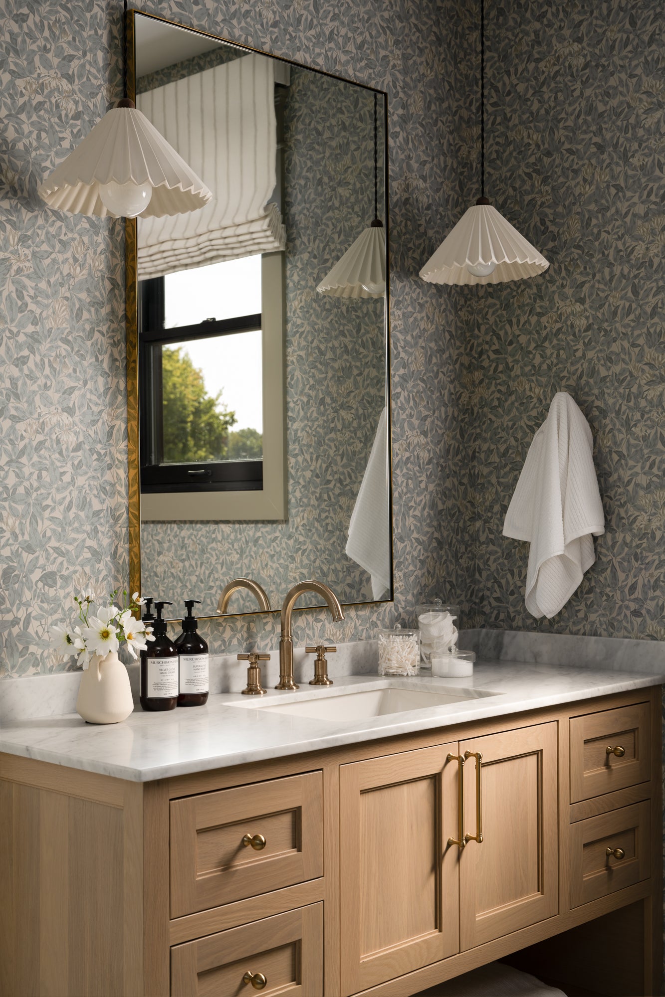 Elegant bathroom with a wooden vanity, marble countertop, gold fixtures, and dual pendant lamps. A large mirror reflects a window and floral wallpaper.
