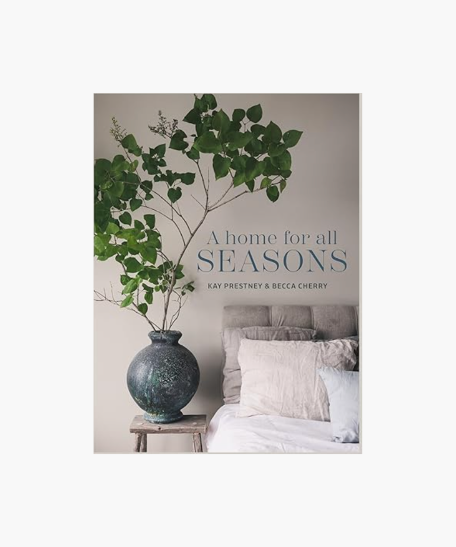 A cozy bedroom scene with a large vase of green leaves on a small wooden stool beside a beige bed. Text reads "A Home for All Seasons" by Kay Prestney & Becca Cherry. | S.W. Home
