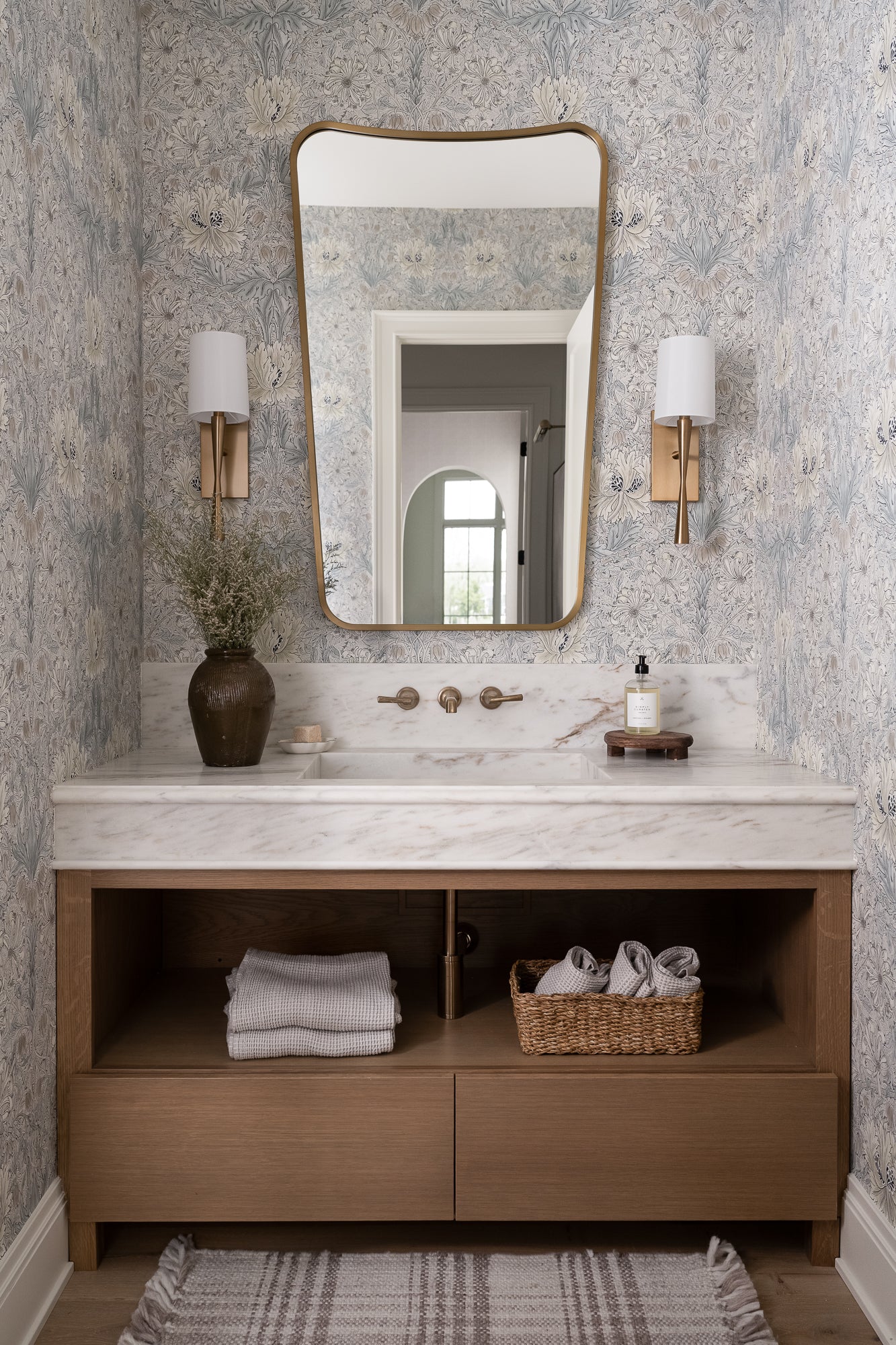 Elegant bathroom with a marble countertop, rectangular mirror, and twin wall sconces. A woven basket and folded towels add warmth to the serene setting. | Wildwood Home Co.