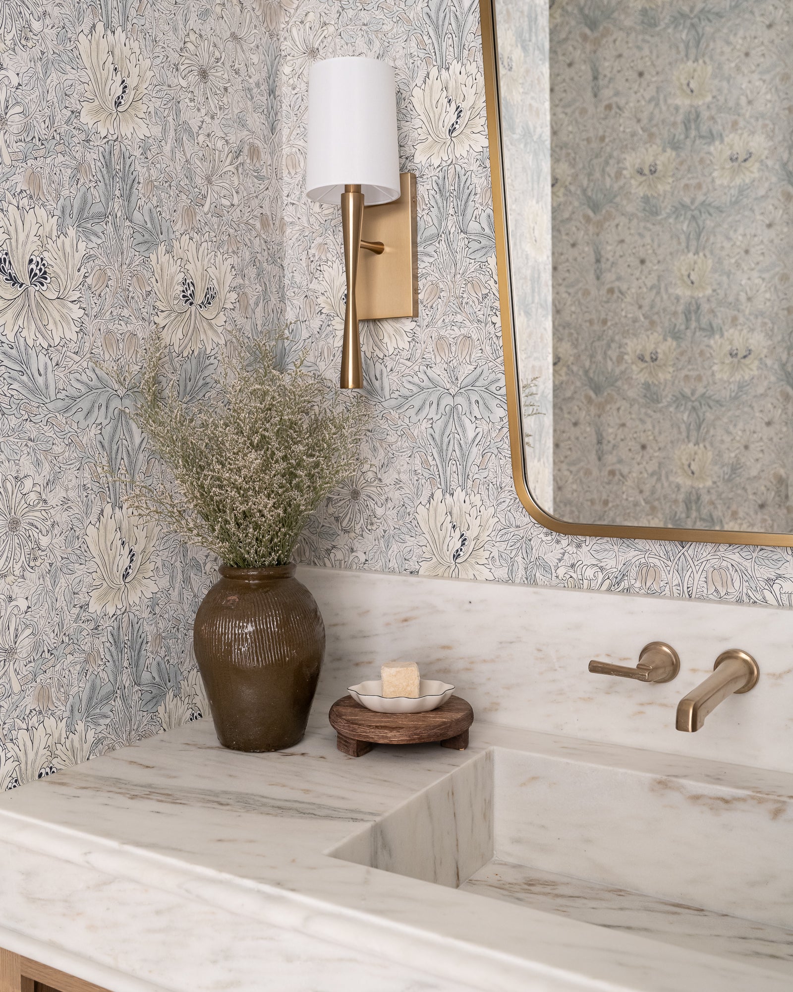 A bathroom features floral wallpaper, a marble sink, brass fixtures, and a rounded mirror. A vase with greenery and soap on a wooden tray add elegance.