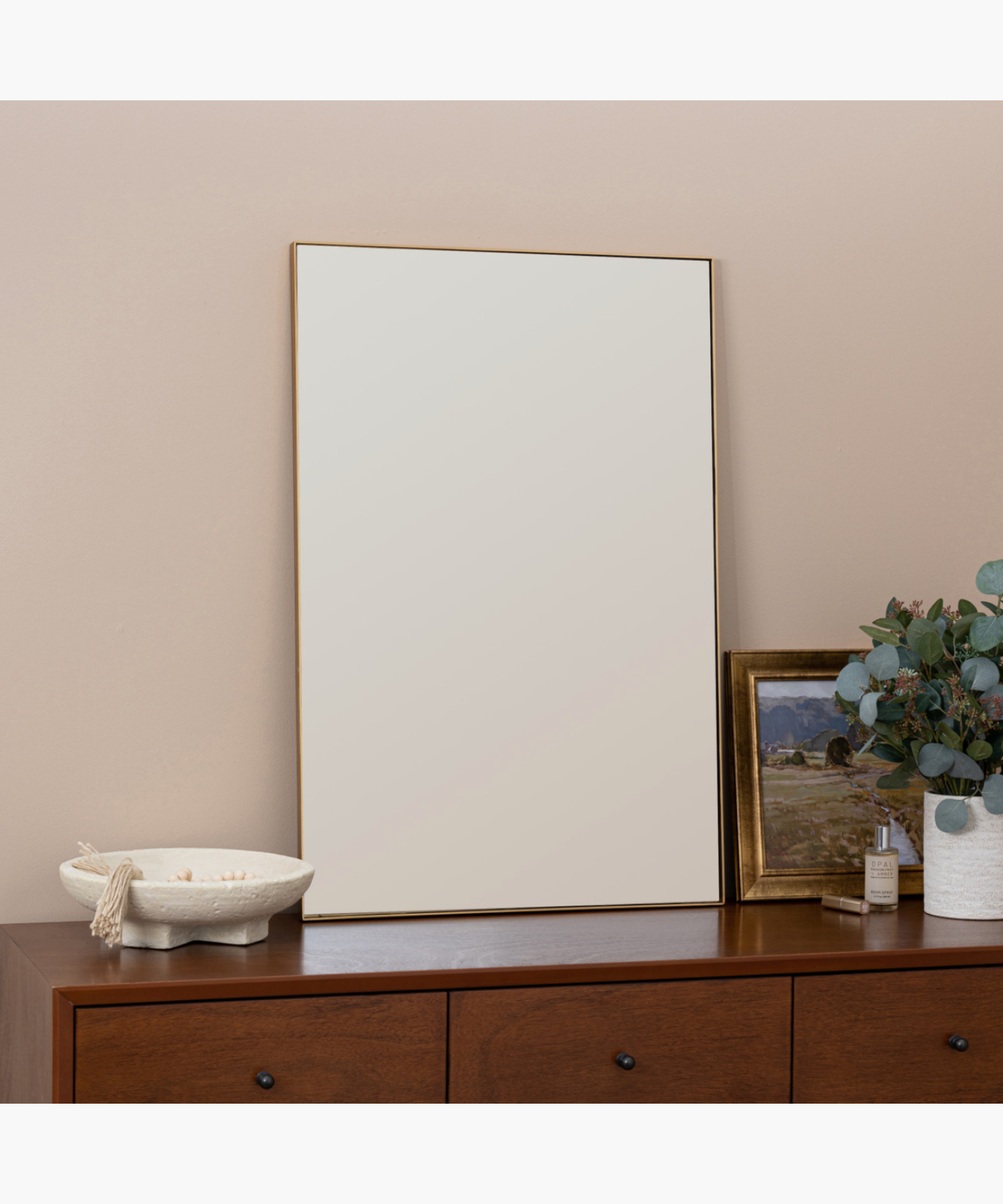 A minimalist living room features a large rectangular mirror on a wooden dresser. Beside it, a vase with eucalyptus, a landscape painting, and a candle. | Wildwood Home Co. 