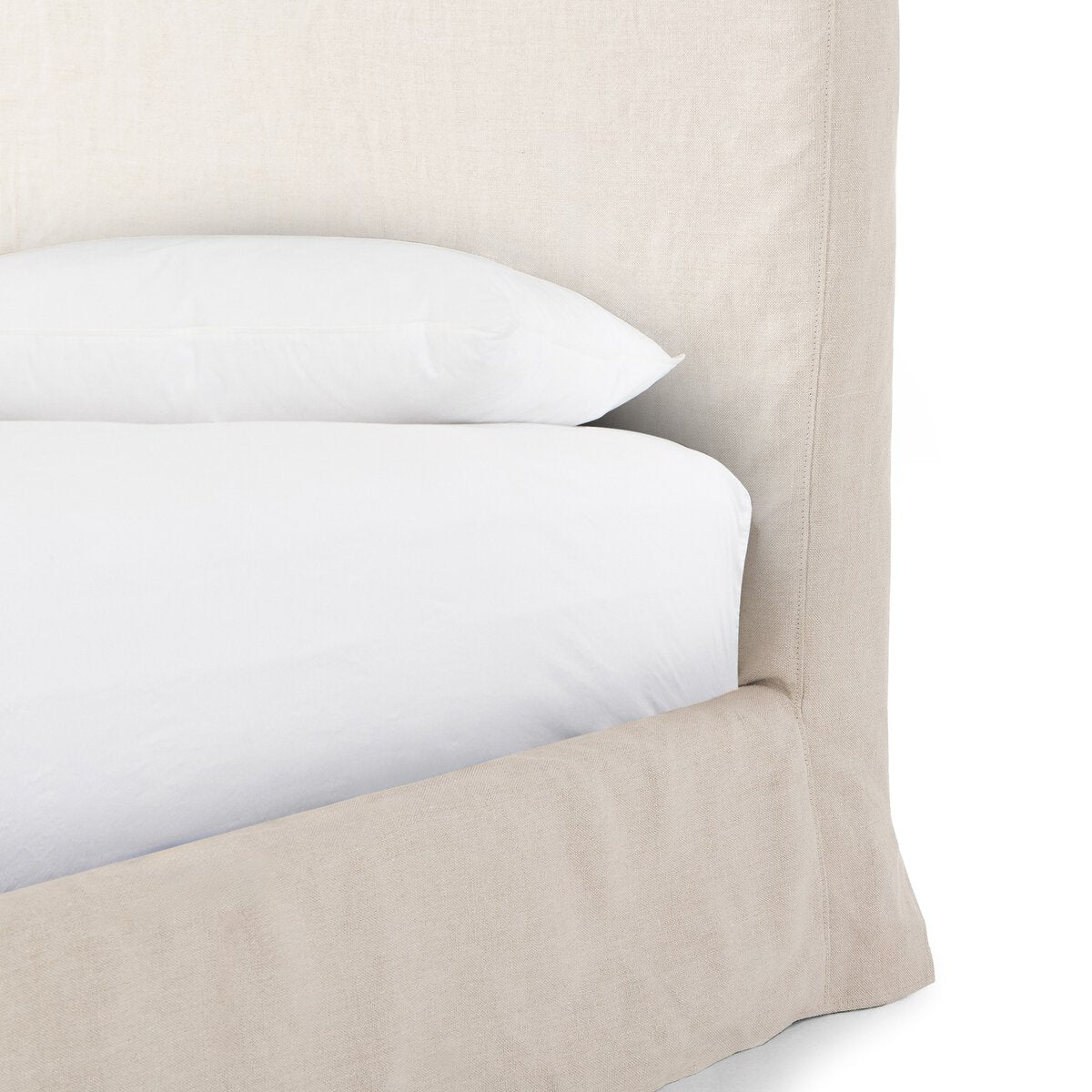 Close up of a beige slipcover bed with white bedding on a white background.