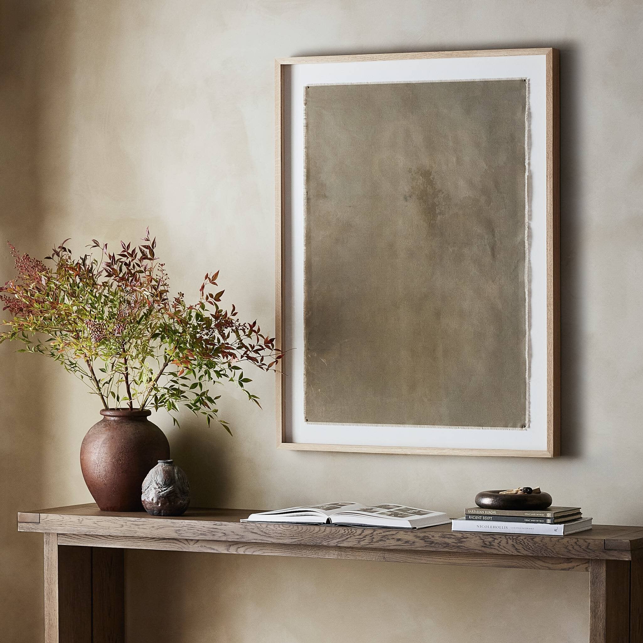 Decorative setup with a vase of flowers, books, and a framed abstract painting on a wooden console against a beige wall. | Wildwood Home Co.