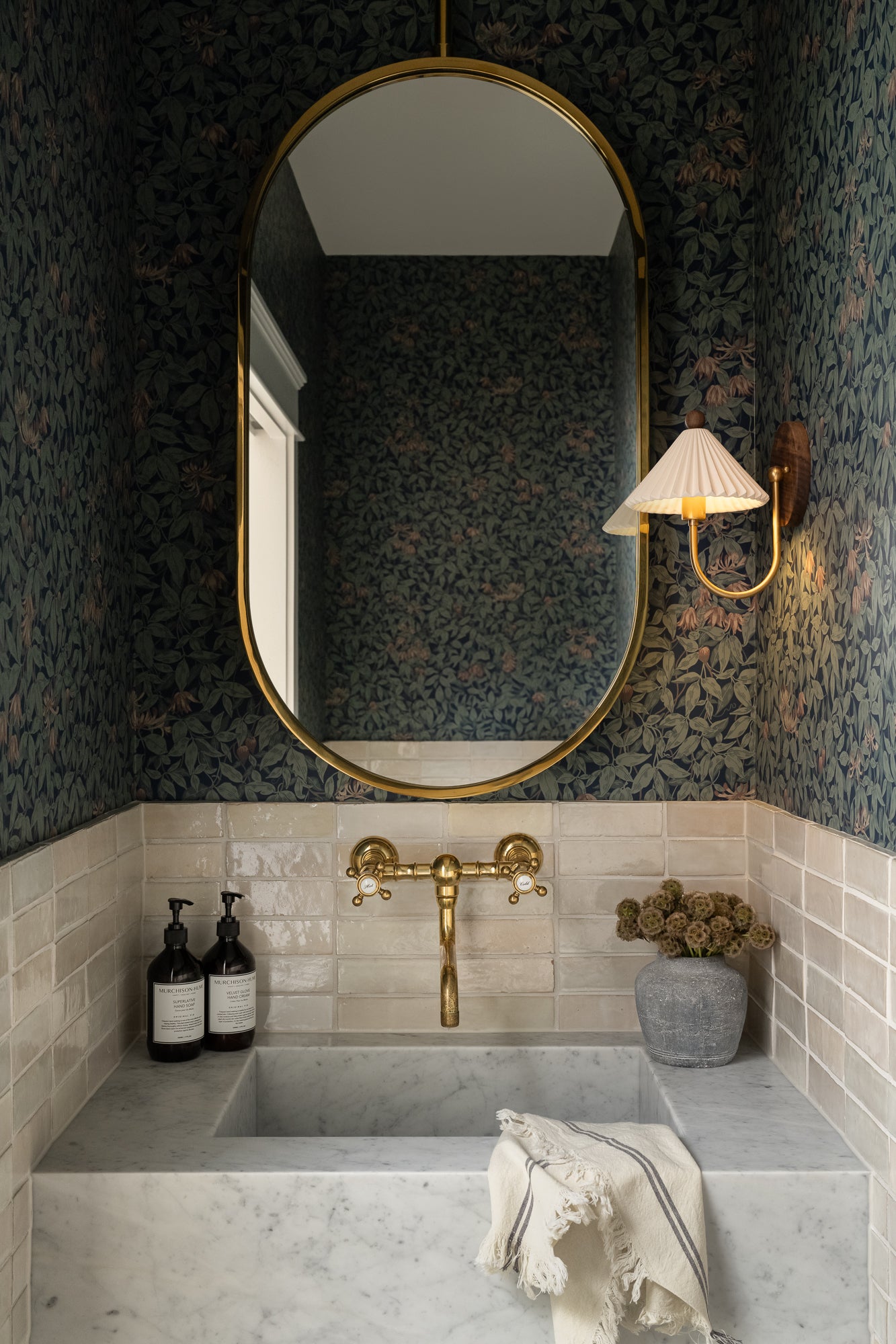Elegant bathroom with floral wallpaper, a gold oval mirror, and brass faucet above a marble sink. A wall sconce and decorative soap bottles add charm. | Wildwood Home Co.