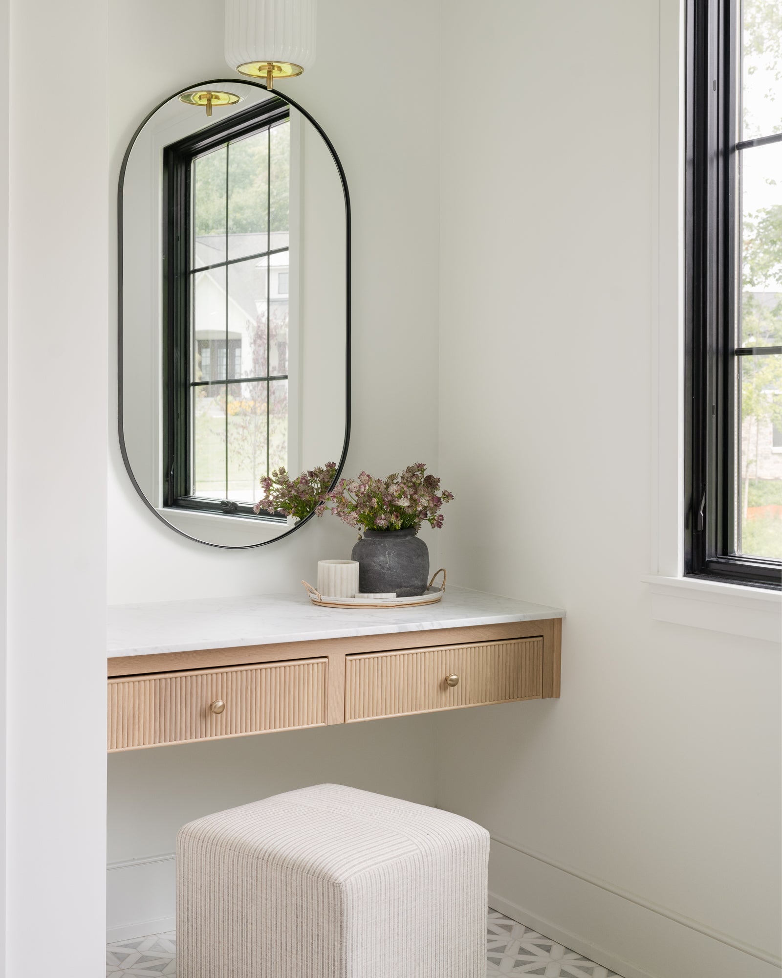 A minimalist vanity area with a large oval mirror, light wood drawers, and a plush, white stool. A vase of flowers adds a touch of nature and elegance. | Wildwood Home Co.