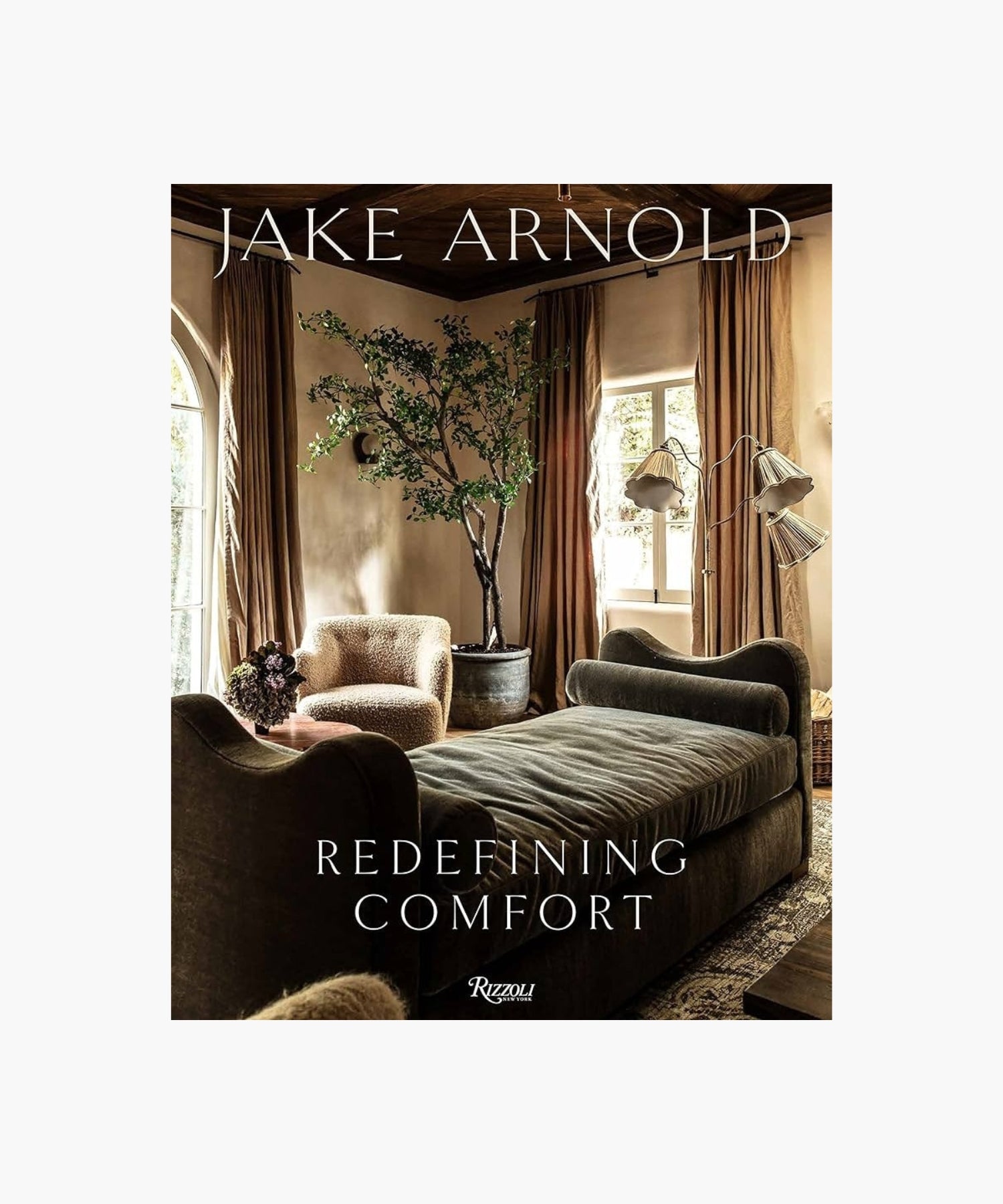 The cover of the book "Redefining Comfort" by Jake Arnold features a cozy living room with plush sofas, an elegant large potted plant, and soft lighting. Earthy tones create a warm, inviting ambiance with a touch of luxury. | S.W. Home