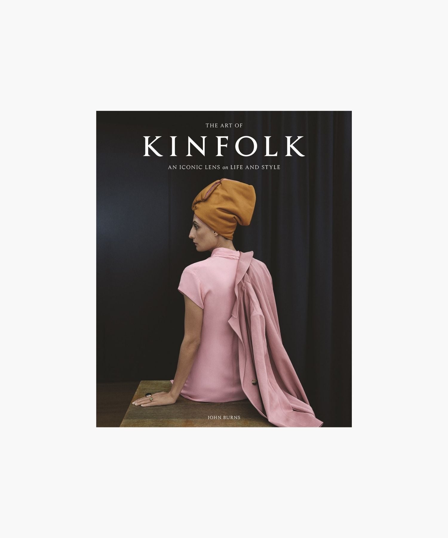 The cover of the book "The Art of Kinfolk" by John Burns features a woman in a pink dress and orange headwrap, sitting elegantly against a dark curtain. The tone is stylish and serene. | S.W. Home