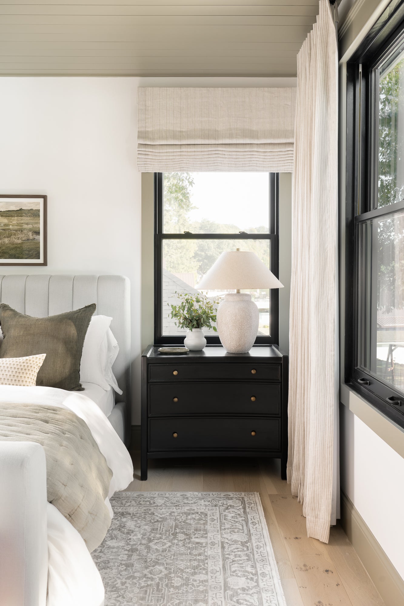 A cozy bedroom with a serene tone features a gray tufted bed, layered pillows, and a beige throw. A dark bedside table holds a ceramic lamp and plant. | SW Home