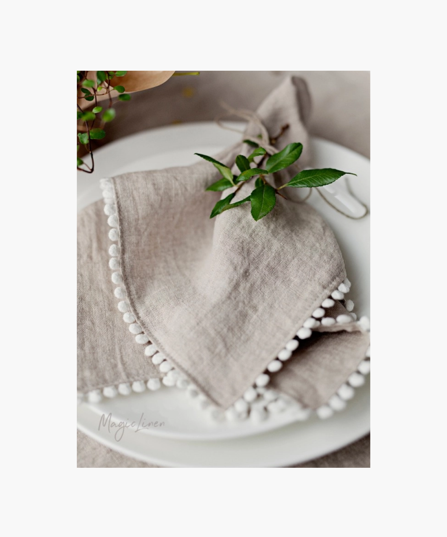 Elegant table setting featuring a delicate beige linen napkin with white pom-pom trim, accented by fresh green leaves on a white plate. | S.W. Home